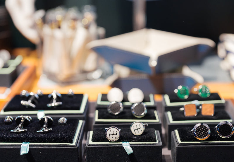 A display case with various pairs of cufflinks arranged neatly on black velvet holders, featuring different shapes, colors, and materials in a well-lit store.