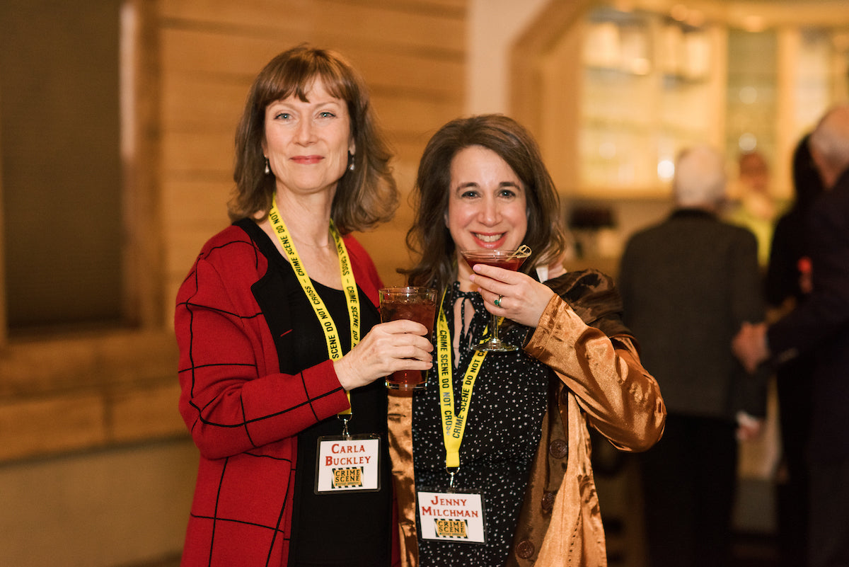 Two women at an event wearing name badges and yellow lanyards, smiling and holding drinks. The woman on the left wears a red jacket, and the woman on the right wears a brown jacket. Other people are blurred in the background.