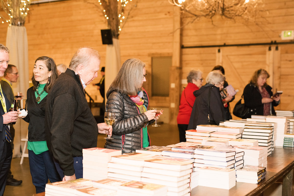 People browse stacks of books on a table at an indoor event. Some are holding drinks, and the room has wooden walls and warm lighting with string lights on wooden beams.