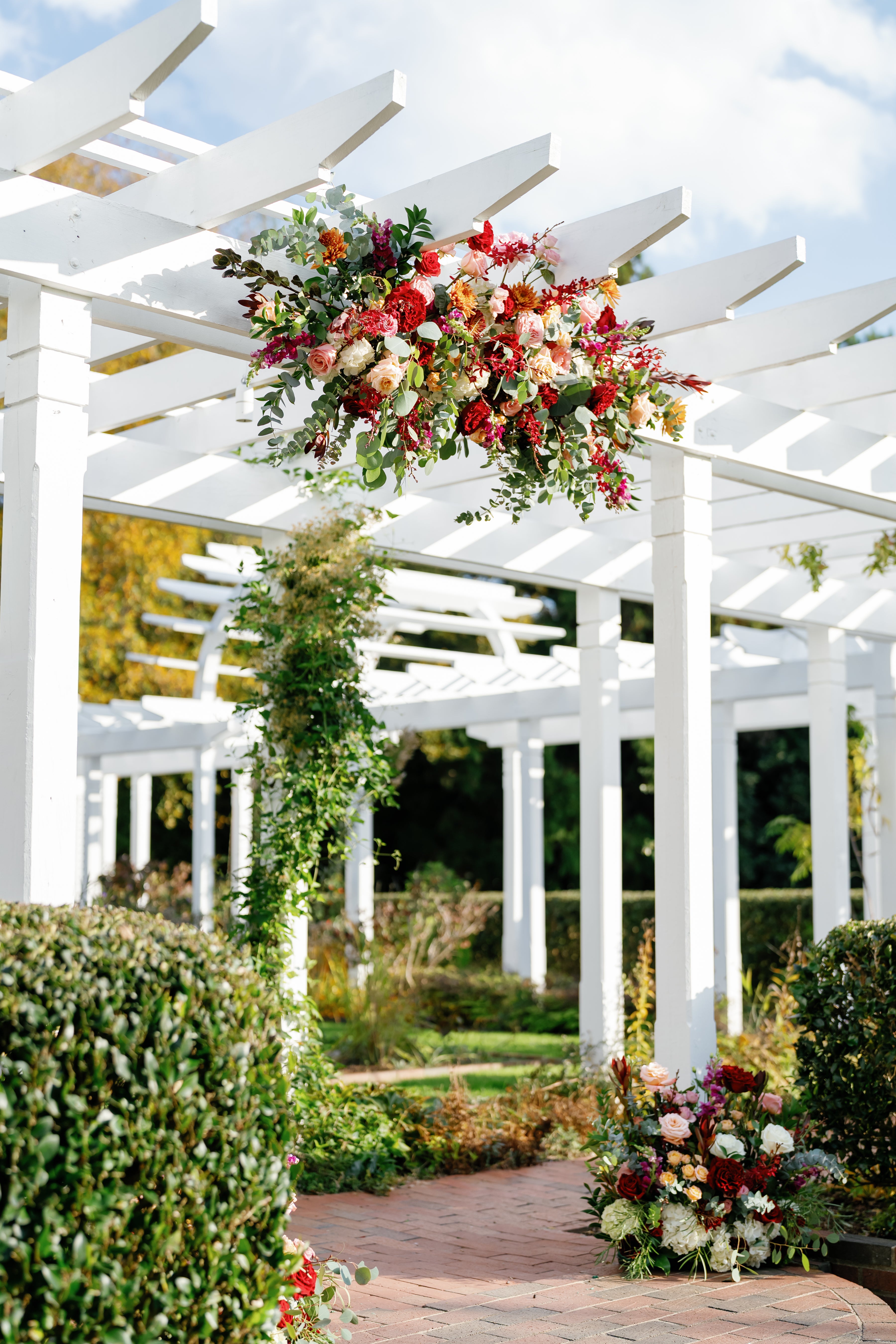A white pergola with lush red and white floral arrangements, green vines, and bushes, set in a sunny garden with a clear blue sky.