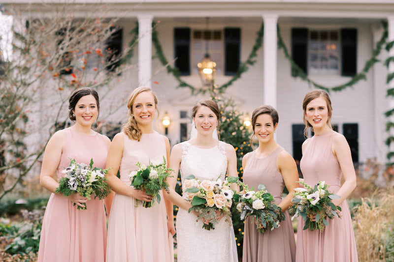 Five women in pink dresses holding floral bouquets in front of a decorated building.