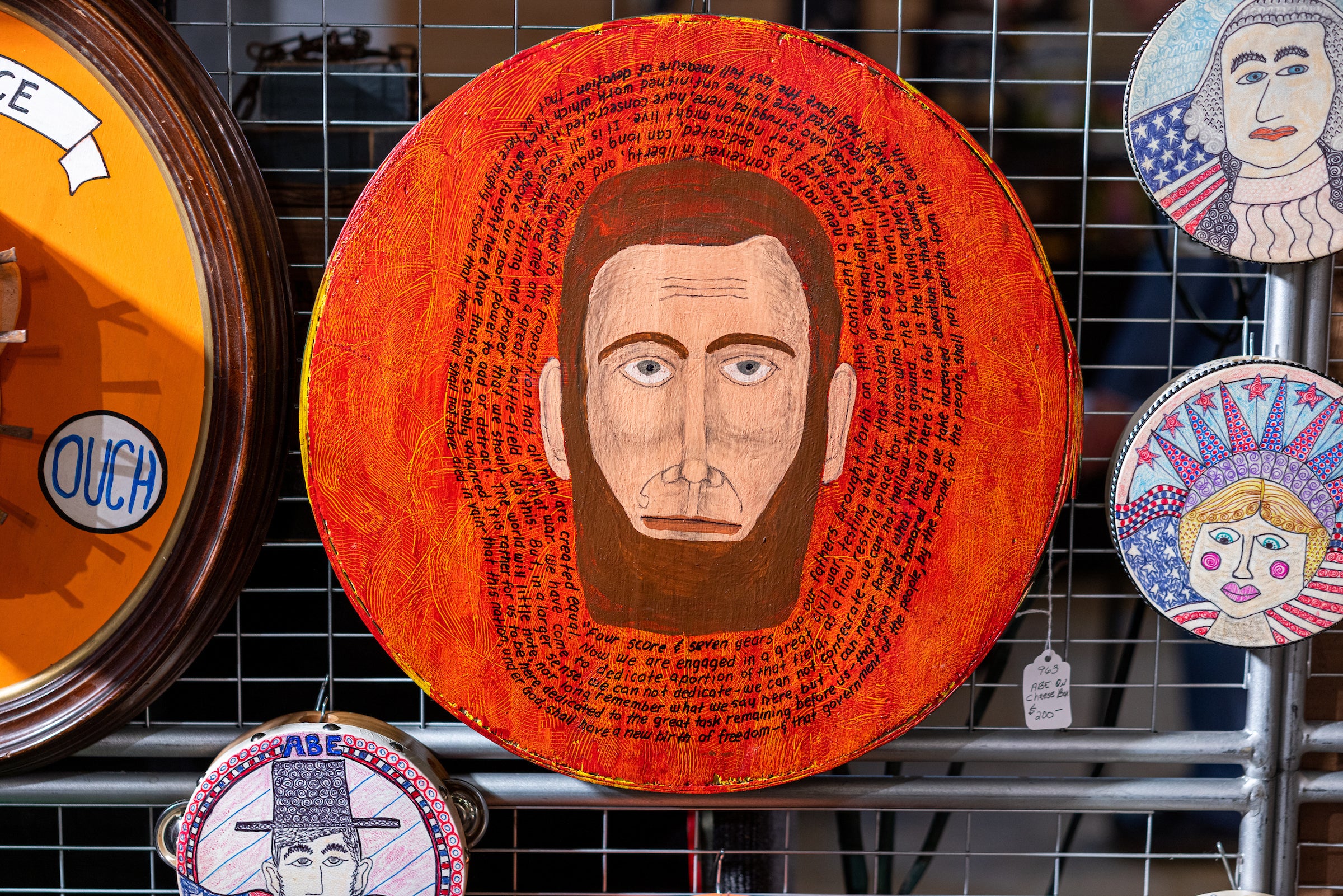 A circular artwork with a bearded man's face in the center, surrounded by handwritten text on a red-orange background, displayed on a wire grid with other colorful portrait art pieces nearby.