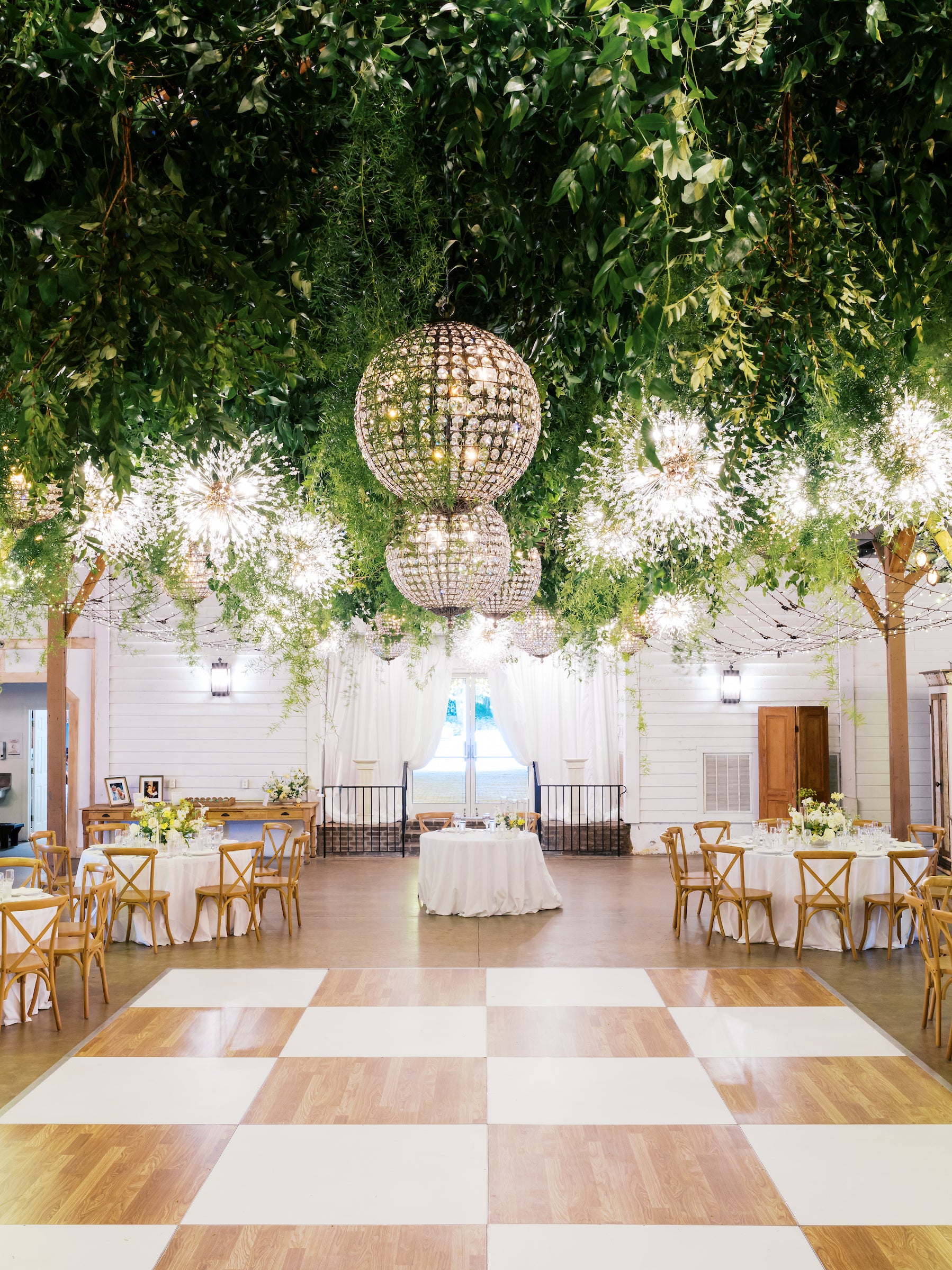 A decorated event hall with a checkered dance floor, round tables with chairs, and lush greenery hanging from the ceiling. Three large chandeliers with crystal ornaments add elegant lighting above the central table.