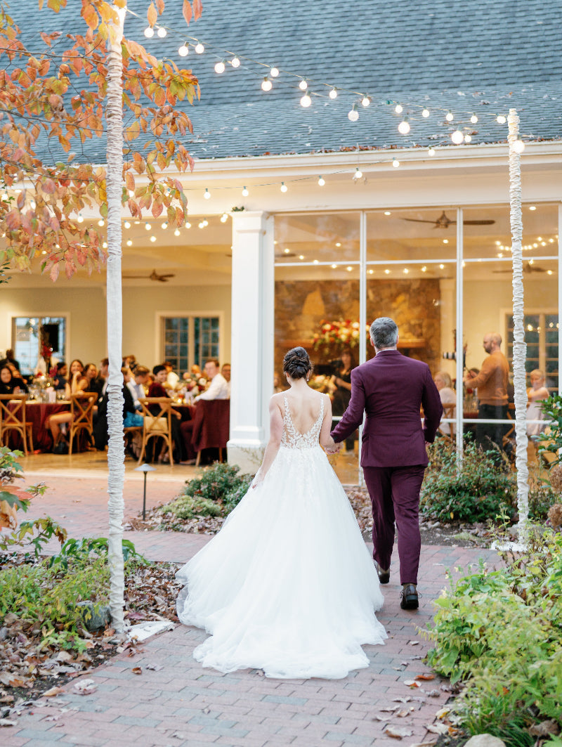A bride in a white gown and a groom in a burgundy suit walk hand in hand toward a warmly lit venue, where guests are seated inside. String lights and autumn trees create a festive, romantic atmosphere.