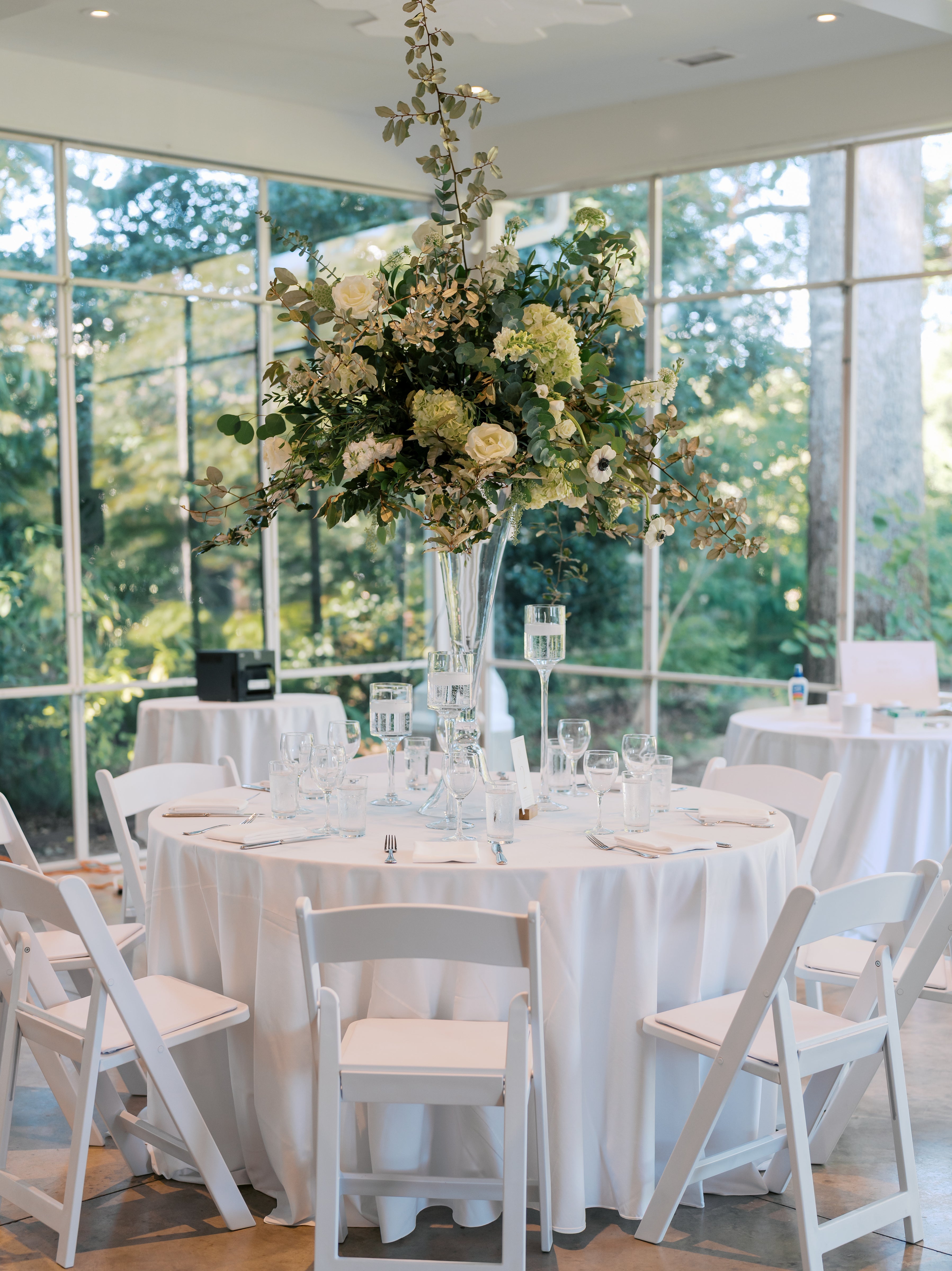 A round table with a white tablecloth is set for a formal event, featuring white chairs and a tall glass vase centerpiece filled with white and green flowers. Large windows reveal lush greenery outside.
