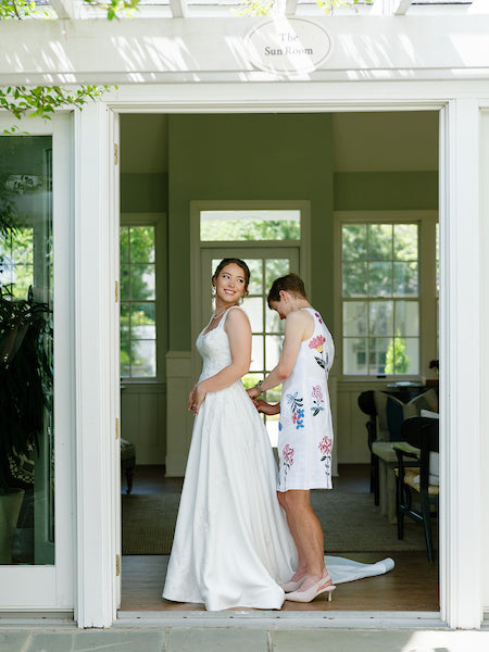 A bride in a white gown stands smiling as another woman in a floral dress helps adjust her dress in a sunlit room, with large windows and greenery visible outside.