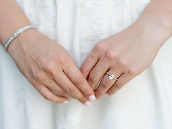 Close-up of a person’s hands in front of a white dress, showing a diamond engagement ring on the left hand and a silver bracelet on the right wrist.