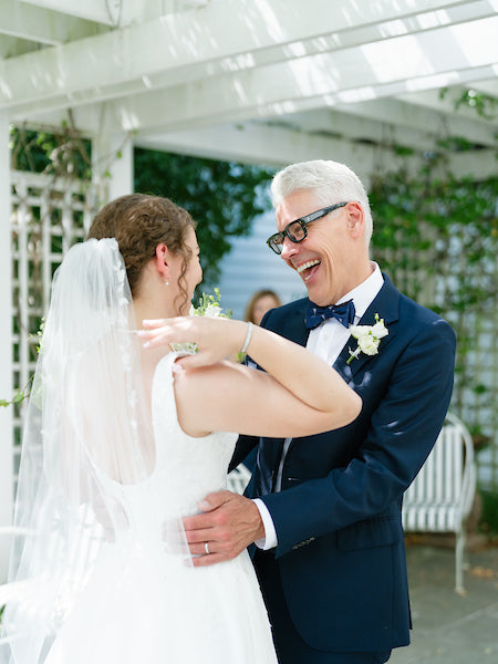 A bride in a white dress and veil shares a joyful moment with an older man in a navy suit and bow tie, both smiling and laughing under a sunlit pergola.
