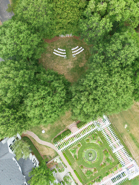 Aerial view of a garden with geometric pathways, greenery, and a circular seating area with white chairs arranged in the middle of a clearing surrounded by trees.