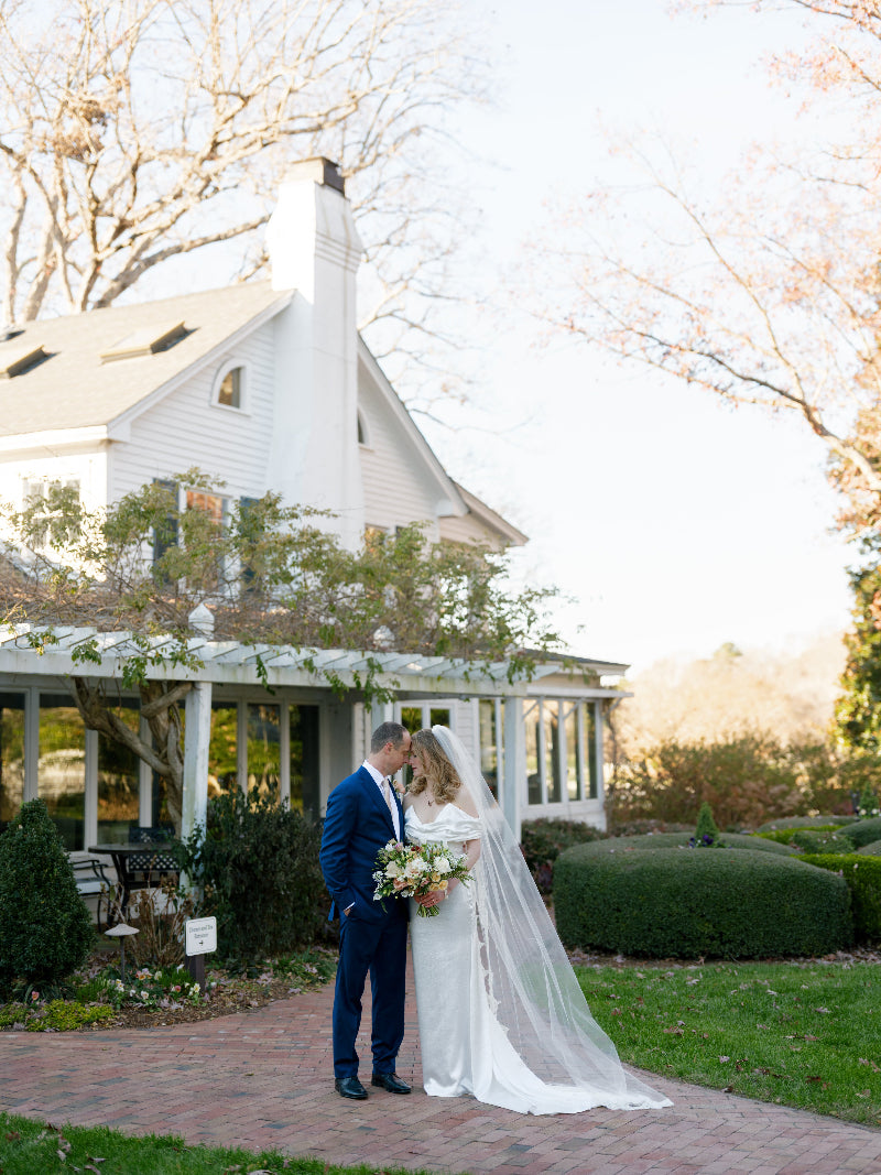 A bride and groom stand closely together on a brick path, with the bride in a white dress and veil holding a bouquet. They are in front of a large white house with a porch, surrounded by trees and greenery.
