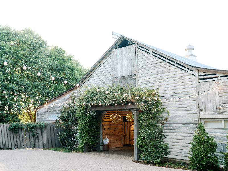 A rustic wooden barn decorated with string lights is surrounded by greenery and trees, with an open entrance leading inside and a paved courtyard in front.