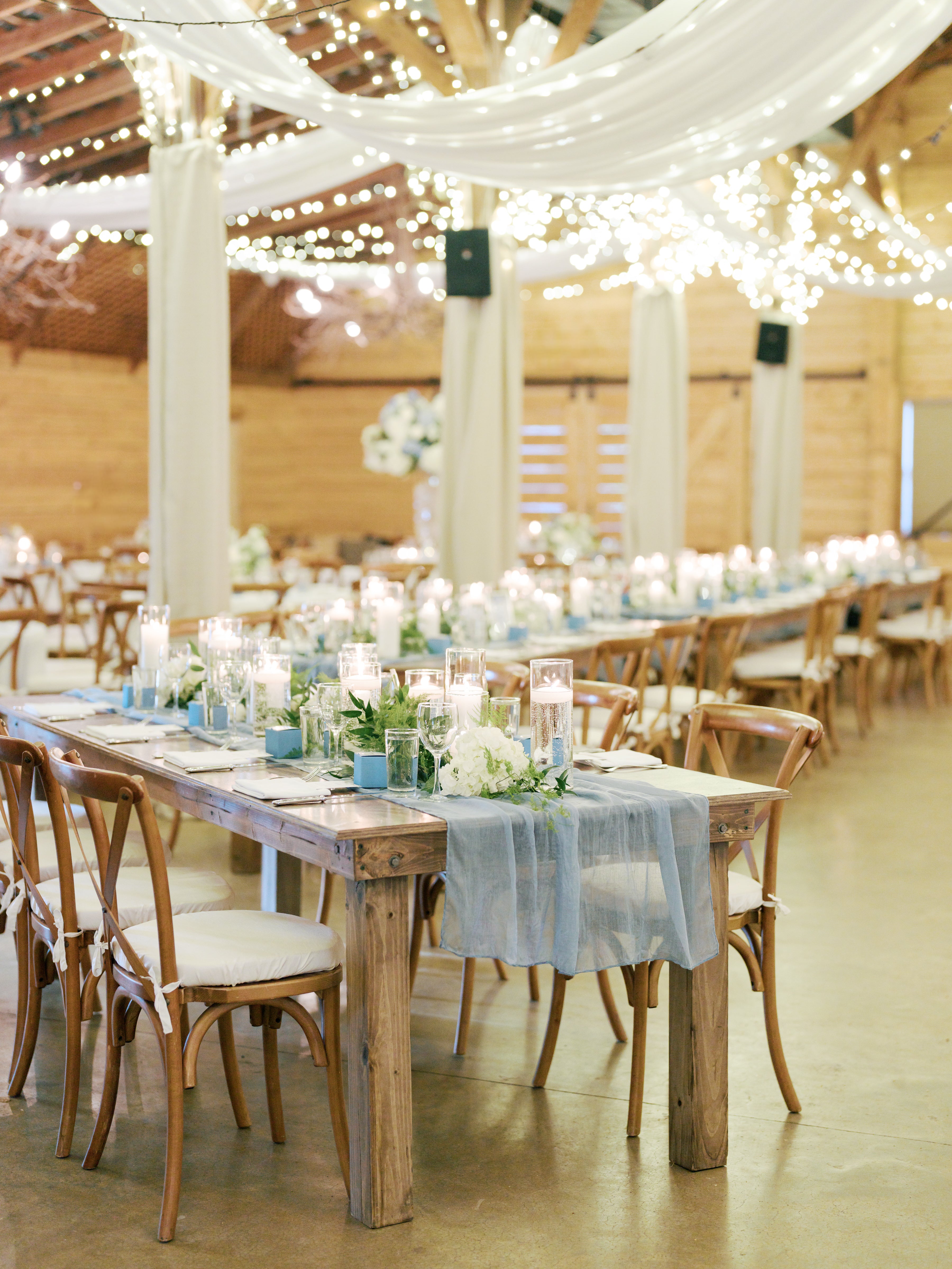 A rustic wedding reception setup features long wooden tables with blue runners, white flowers, candles, and wooden chairs. String lights and draped fabric hang from the ceiling, creating a warm, romantic atmosphere.