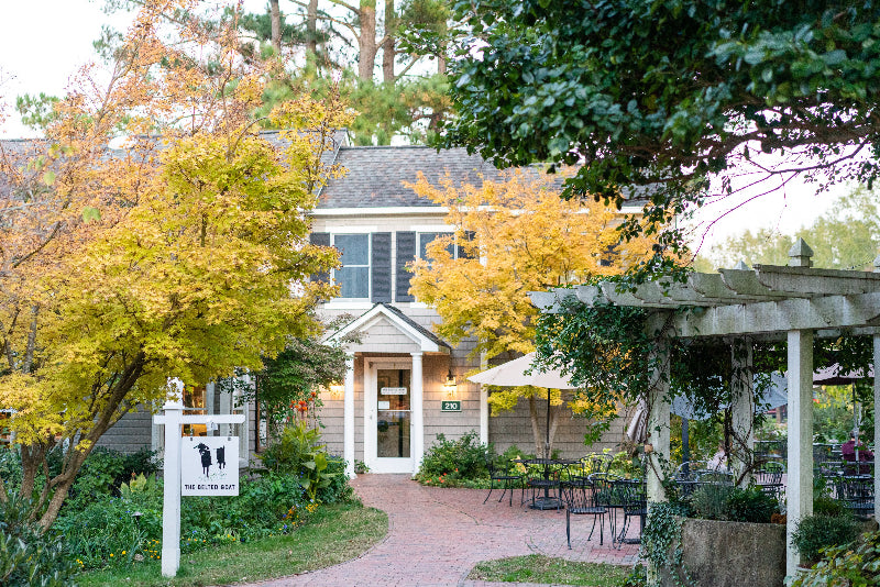 A charming house with yellow autumn trees and lush greenery, featuring outdoor seating, a white sign with silhouettes, and a brick walkway leading to the front entrance.