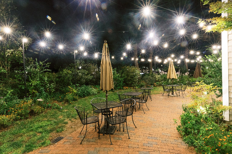 Outdoor patio at night with empty metal tables and chairs, closed umbrellas, brick flooring, surrounding greenery, and string lights creating a warm, inviting atmosphere.