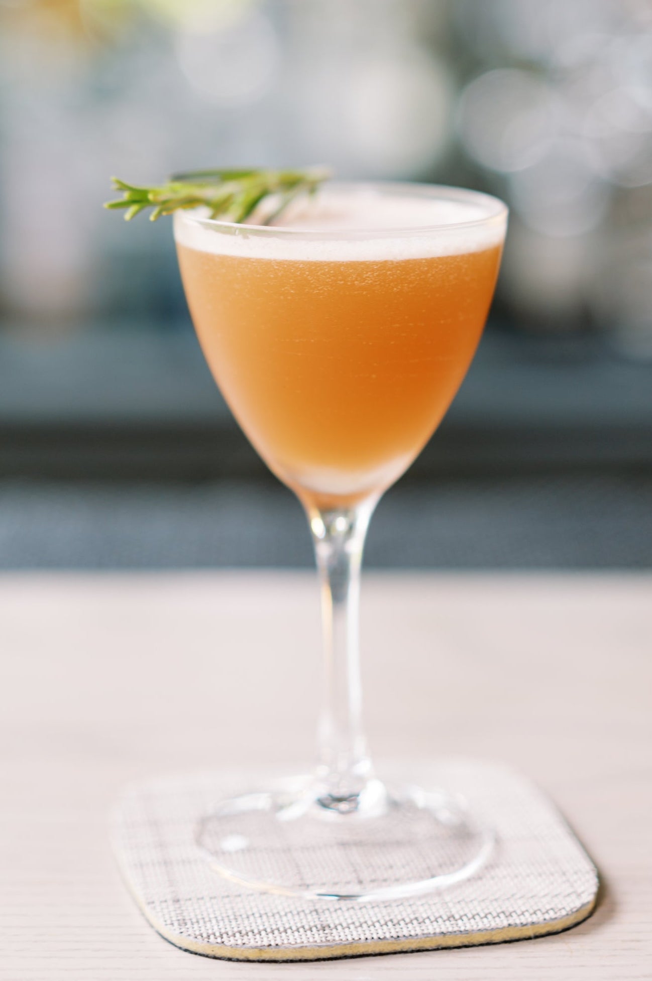 A frothy orange cocktail is served in a stemmed glass, garnished with a sprig of rosemary, and placed on a textured square coaster with a blurred background.