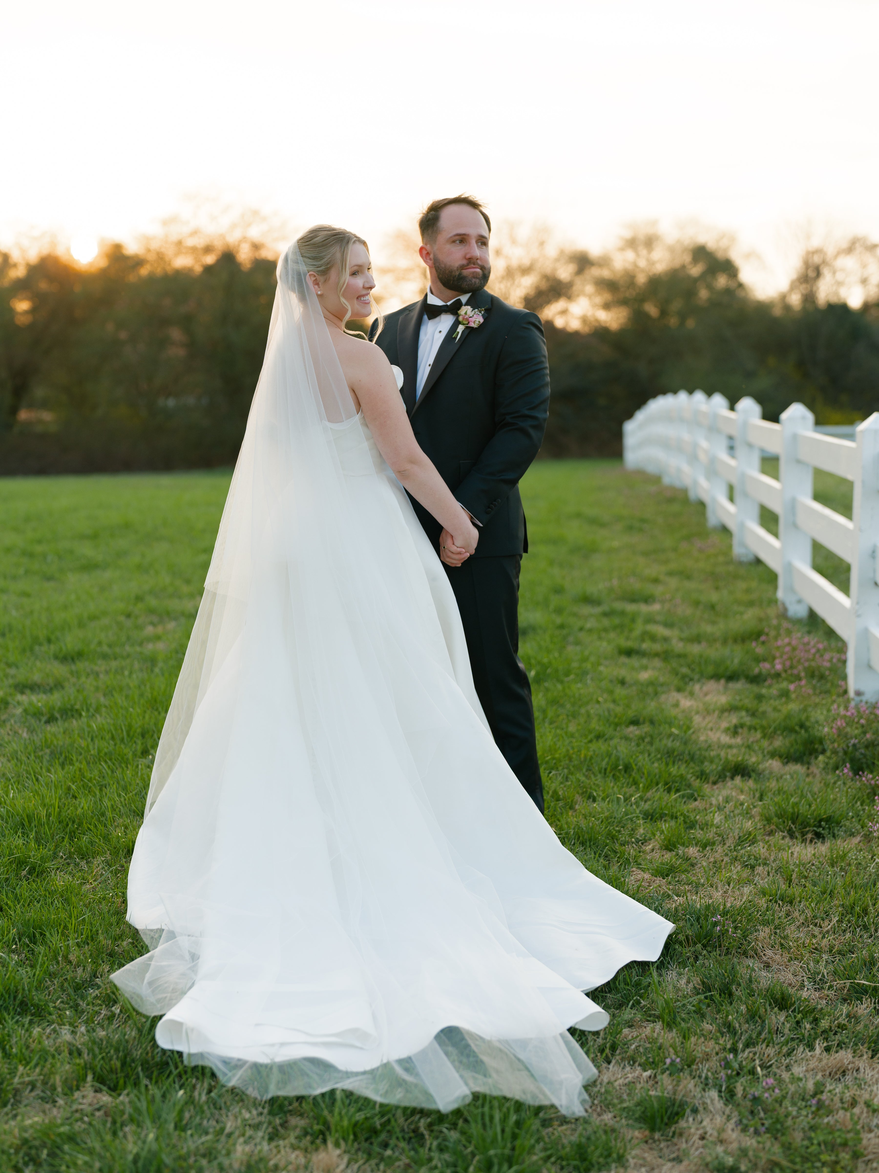 A bride in a white dress and veil stands holding hands with a groom in a black tuxedo on a grassy field near a white fence at sunset.
