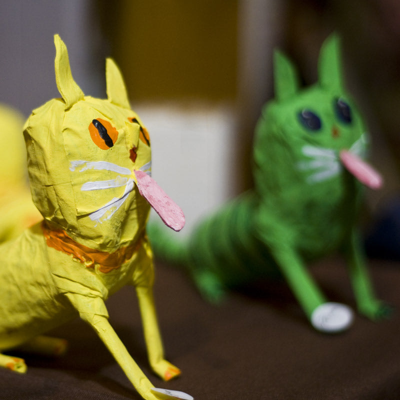 Two colorful paper mache cat sculptures, one yellow and one green, both with large eyes, white whiskers, and long pink tongues sticking out, are displayed on a brown surface.