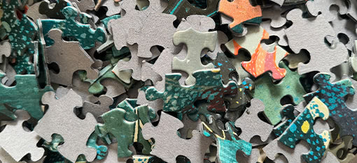 A close-up view of a scattered pile of jigsaw puzzle pieces, showing various colors and patterns including green, gray, and orange. The pieces are randomly arranged and unassembled.