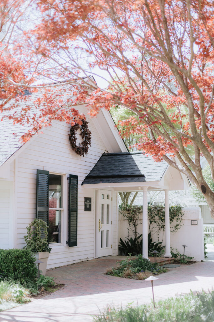 A charming white cottage with black shutters and a wreath on the gable, surrounded by blooming red-leaved trees and lush greenery, sits under a bright, sunlit sky.