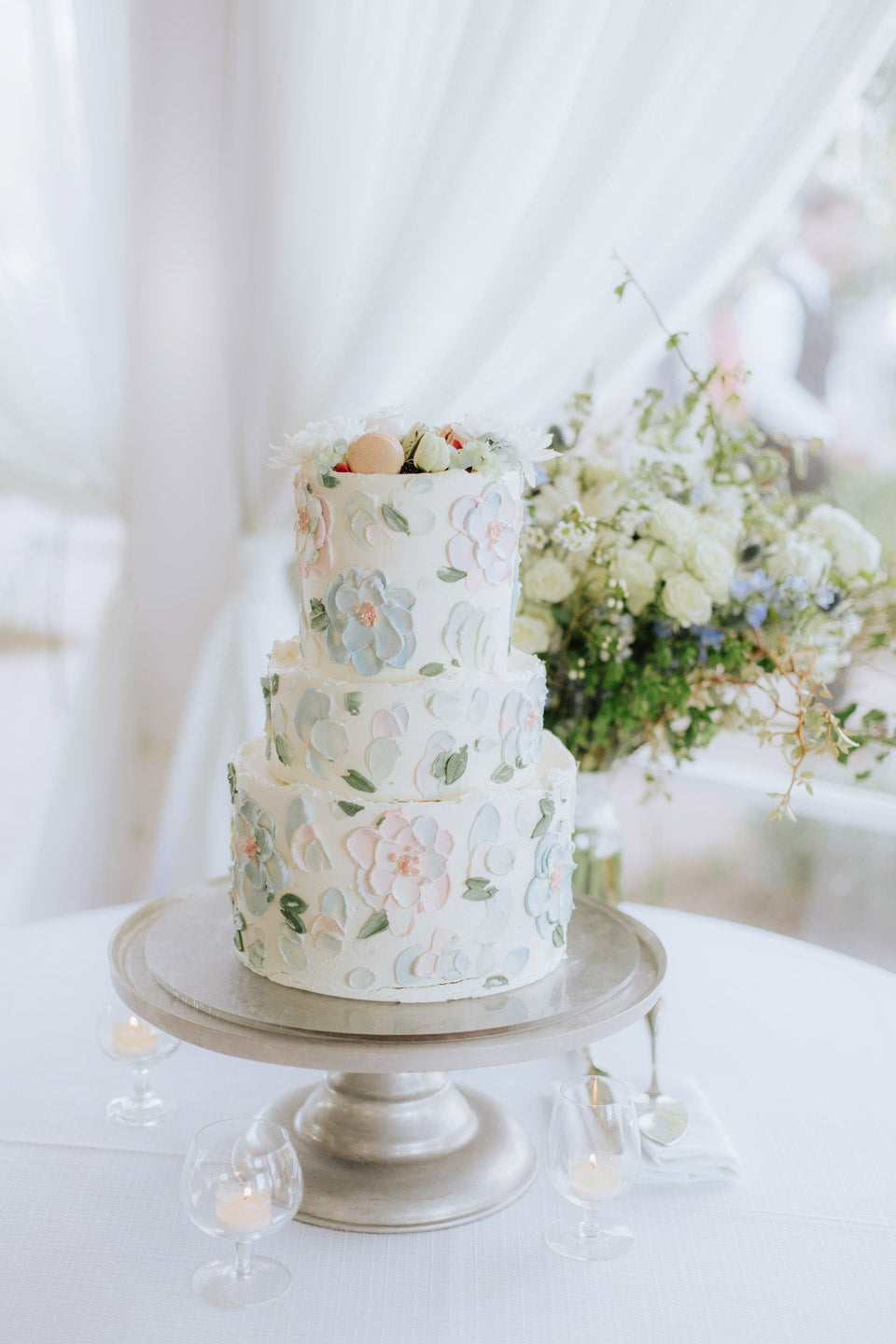 A three-tiered white cake with pastel floral icing sits on a silver stand. The cake is decorated with fresh flowers on top. In the background, there is a bouquet of white flowers and greenery.