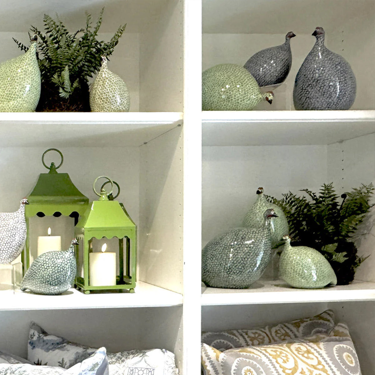 White shelves display decorative ceramic guinea fowl, green lanterns with candles, small potted ferns, and patterned pillows in neutral and yellow tones. The arrangement is neat and visually balanced.