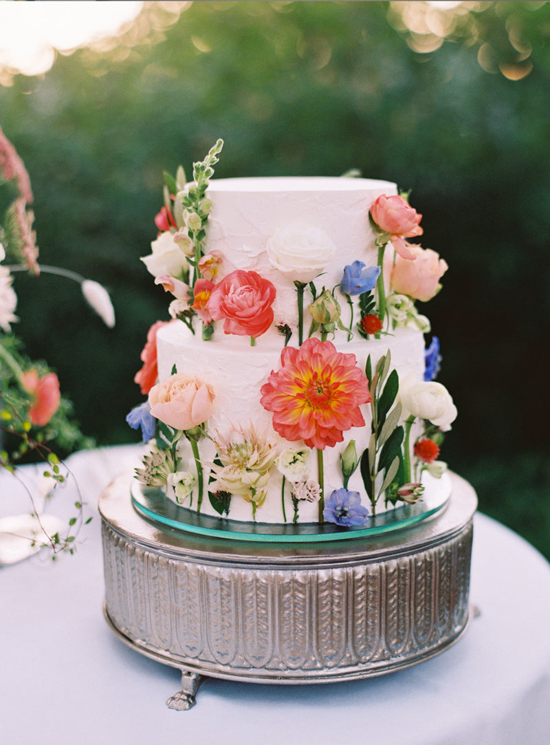 A two-tiered white cake decorated with vibrant fresh flowers in pink, red, white, blue, and orange, displayed on an ornate silver cake stand outdoors.