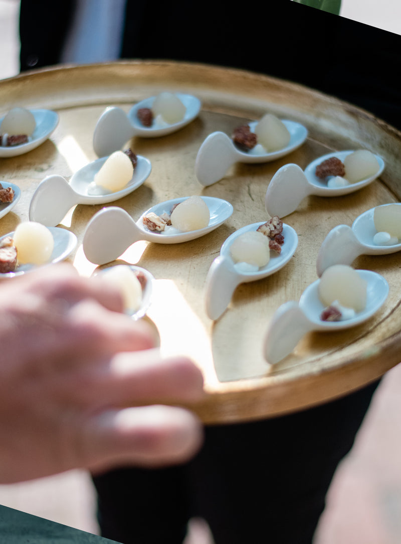 A wooden tray holds several small white ceramic spoons, each with a round bite-sized food item and a garnish, as a hand reaches toward the tray.