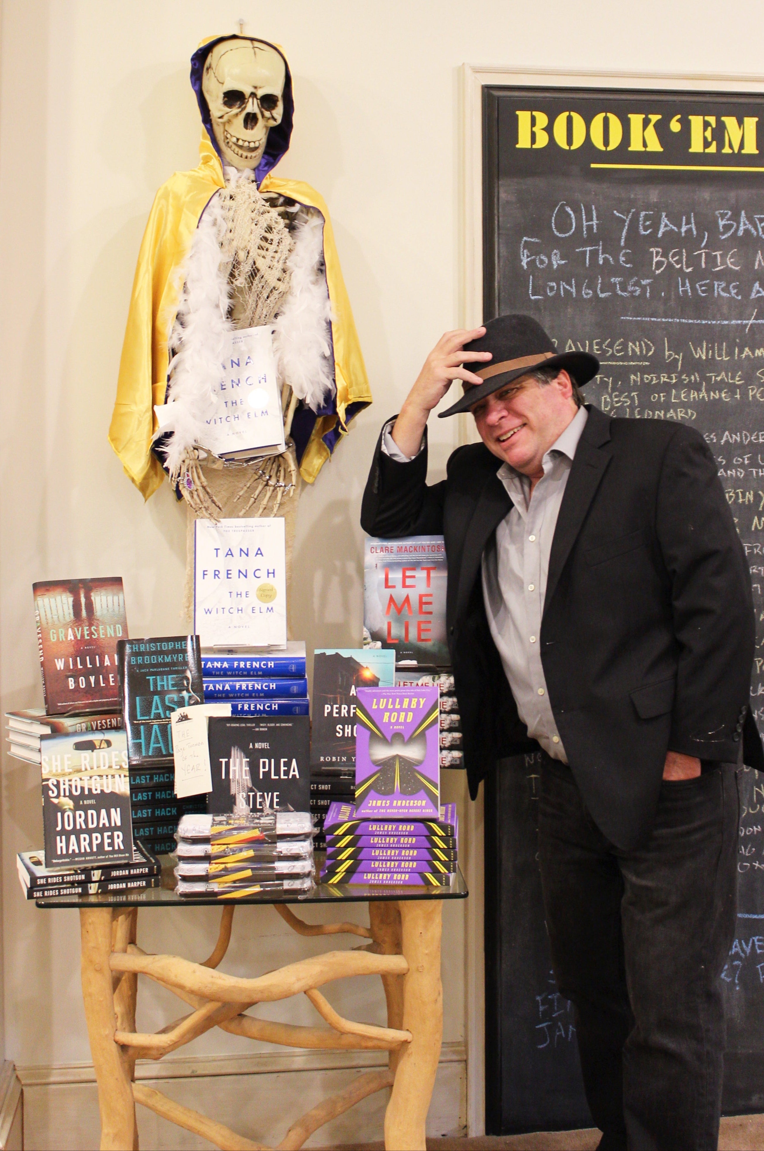 A man in a hat poses and tips his hat next to a table stacked with books. Behind him stands a skeleton draped in a cape and feather boa. A chalkboard and “BOOK’EM” sign are visible in the background.