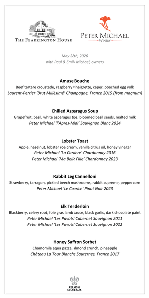 A man in a straw hat and a woman in a denim jacket smile together outdoors by a wooden bench with a stag carving—an inviting scene evoking the Peter Michael Wine Dinner by FEARRINGTON VILLAGE, surrounded by tall trees and greenery.