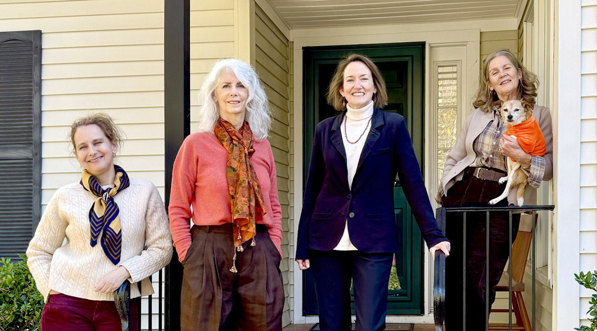 Four women pose on a brick front porch with a green door. One woman holds a small dog wearing an orange sweater. They are dressed in fall or winter clothing and are smiling at the camera.