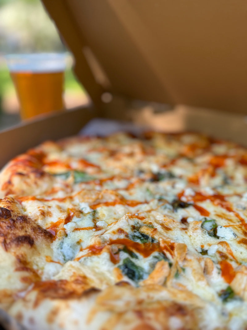 A close-up of a pizza topped with cheese, spinach, and hot sauce in an open pizza box, with a blurred glass of beer in the background.