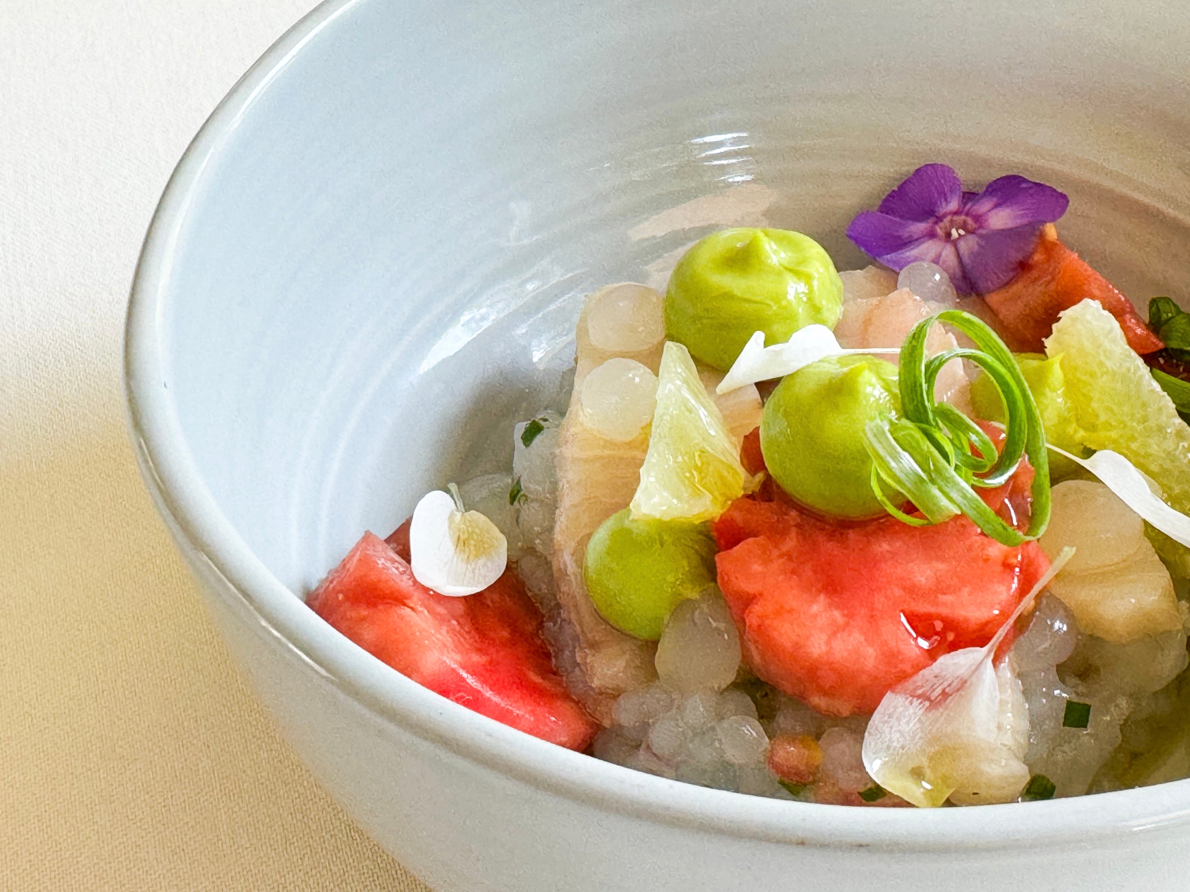 A close-up of a white bowl containing a colorful, gourmet dish with green mousse, translucent pearls, red fruit slices, edible flowers, and garnishes arranged artfully on a light surface.