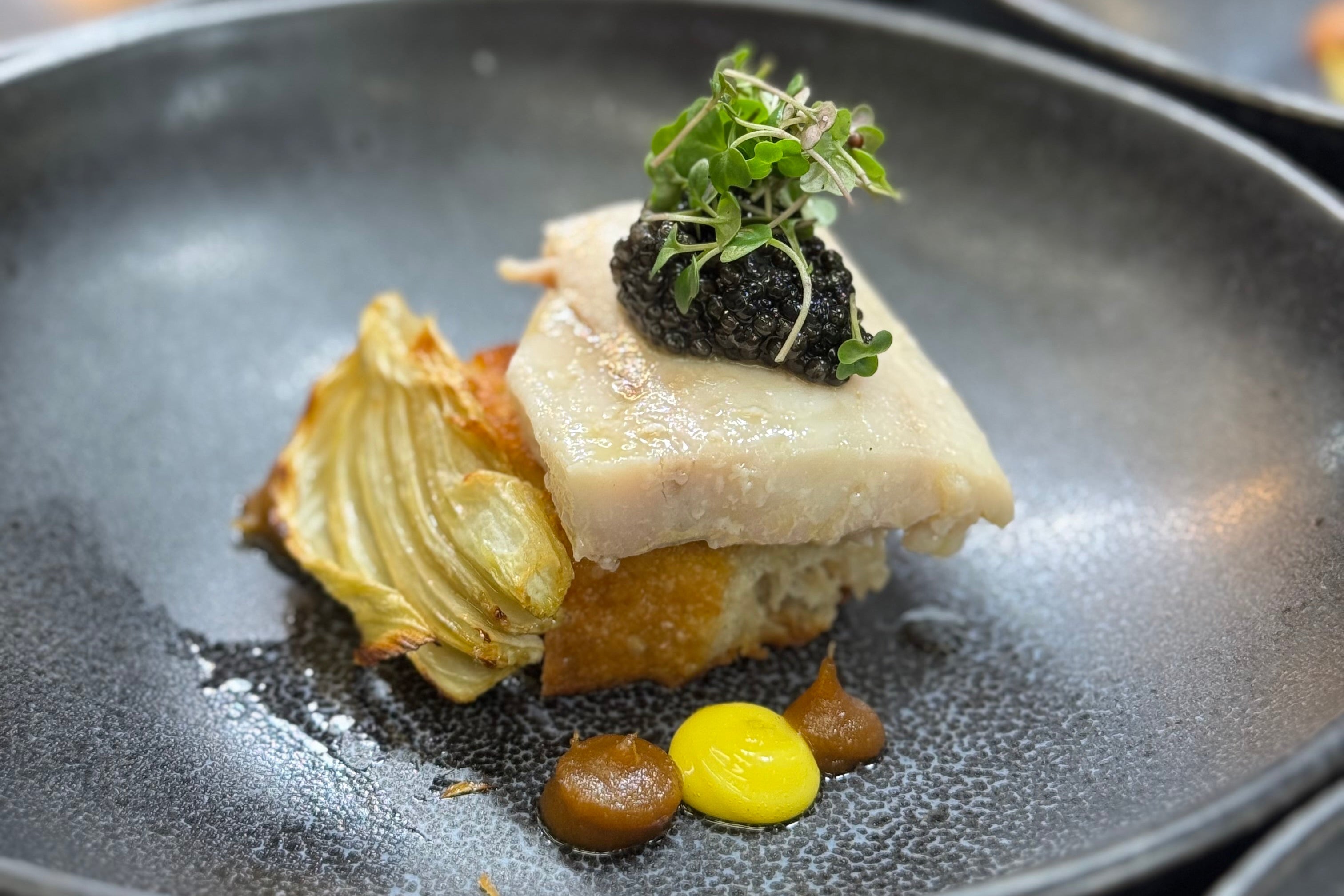 A gourmet dish featuring a piece of white fish on toasted bread, topped with caviar and microgreens, accompanied by roasted fennel and dollops of brown and yellow sauces on a dark plate.