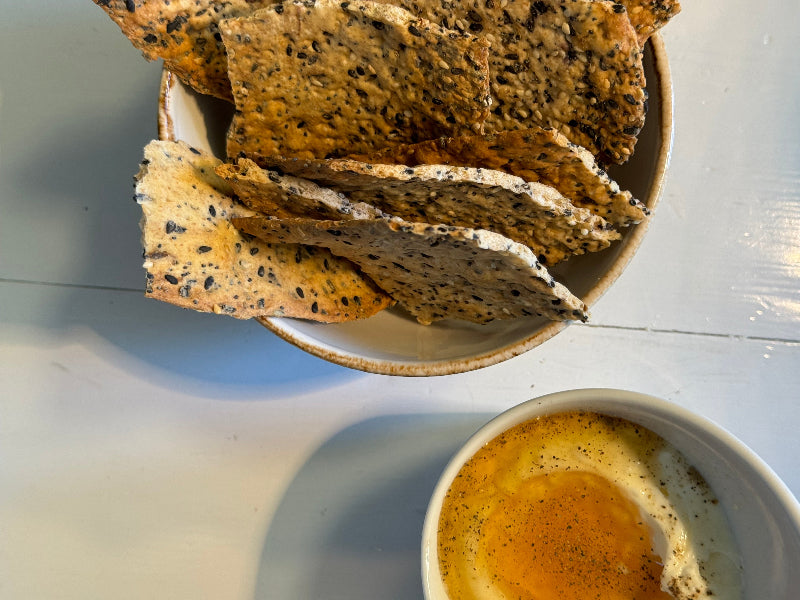 A bowl of seeded crispbread crackers sits next to a small bowl of soft-cooked eggs with runny yolk, topped with black pepper, on a light-colored table.