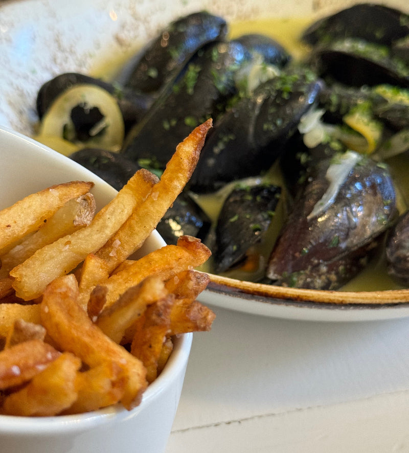 A close-up of crispy golden French fries in a white bowl next to a plate of steamed mussels in a creamy sauce, garnished with chopped herbs.