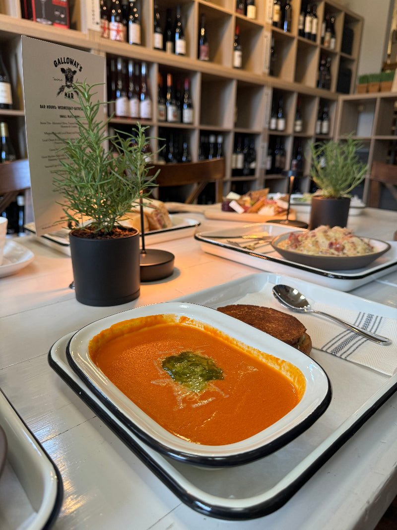A bowl of tomato soup with green garnish sits on a white tray beside a grilled sandwich in a cozy restaurant, with shelves of wine bottles and small potted plants on the tables in the background.
