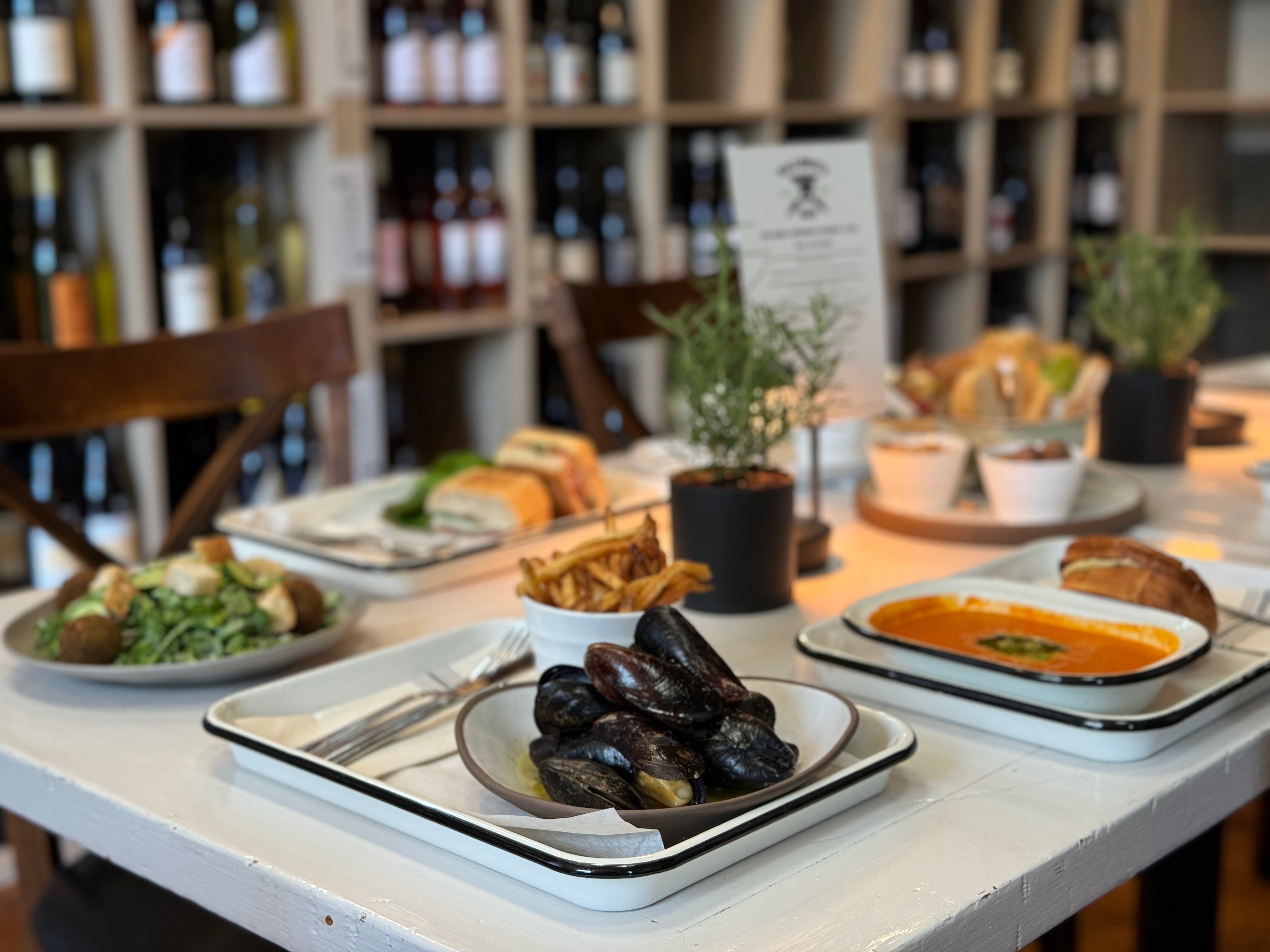 A table set with plates of mussels, soup, salad, French fries, sandwiches, and bread, with shelves of wine bottles in the background and small potted plants as table decor.