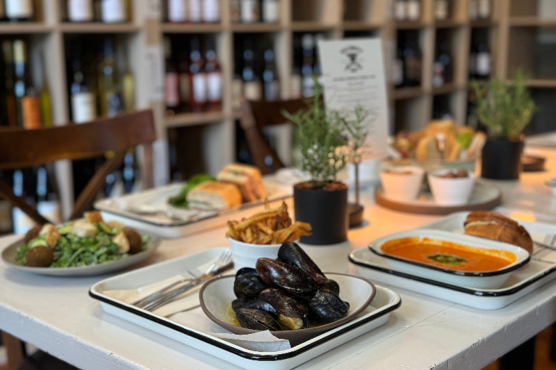 A table set with plates of mussels, fries, salad, soup, and sandwiches in a cozy restaurant with wine bottles displayed on shelves in the background.