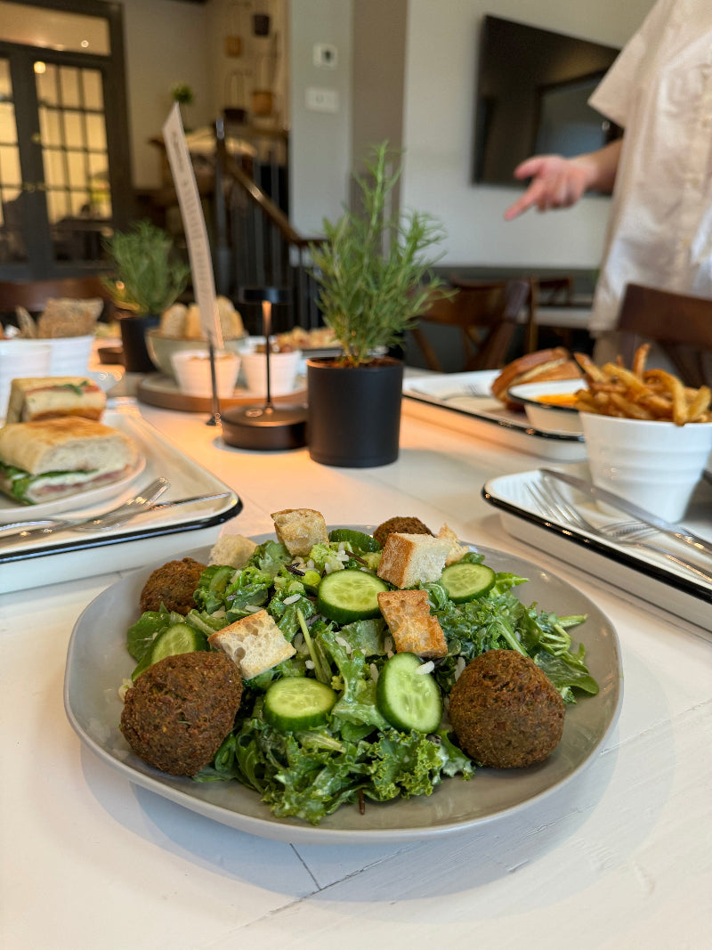 A plate of mixed greens topped with falafel, sliced cucumbers, and croutons sits on a table with sandwiches, fries, and a small potted plant in the background. A person stands nearby, gesturing with their hand.