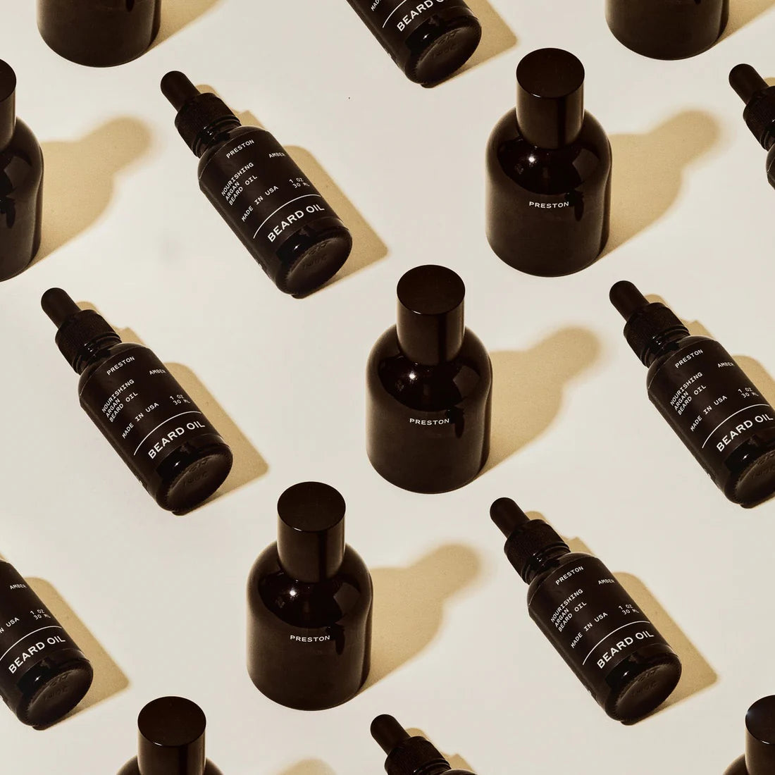 Several black bottles of beard oil and dropper bottles are arranged in a repeating, diagonal pattern on a beige background, creating a modern and visually appealing flat lay composition.