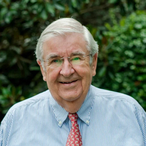 An older man with gray hair and glasses is smiling outdoors. He is wearing a light blue striped shirt and a red tie with a pattern. Green foliage is blurred in the background.