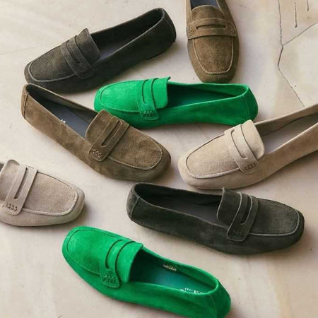 A collection of suede loafers in various colors including olive green, bright green, and beige arranged on a light surface.