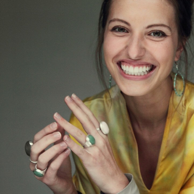 A woman with long brown hair smiles widely, showing her teeth. She wears a yellow patterned top, hoop earrings, and several large, stone rings on her fingers. Her hands are raised and close to her face.