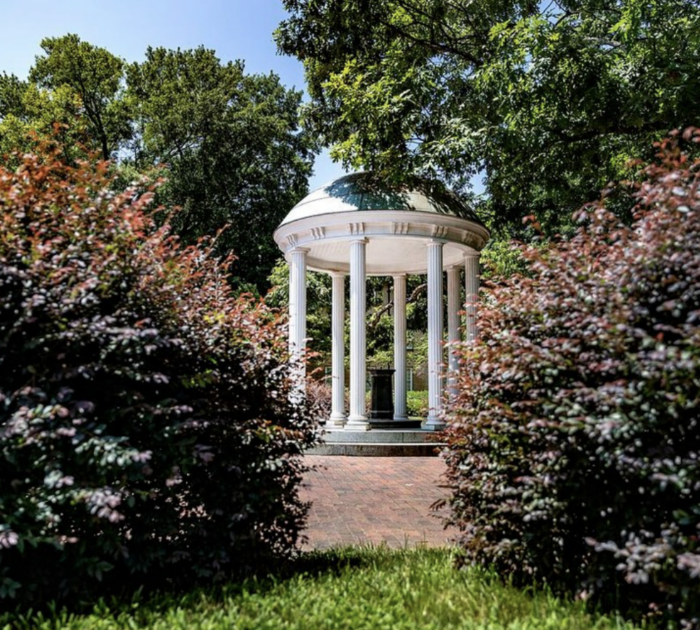 A white, domed gazebo with tall columns stands surrounded by green trees and bushes on a sunny day, with a brick path leading up to it.