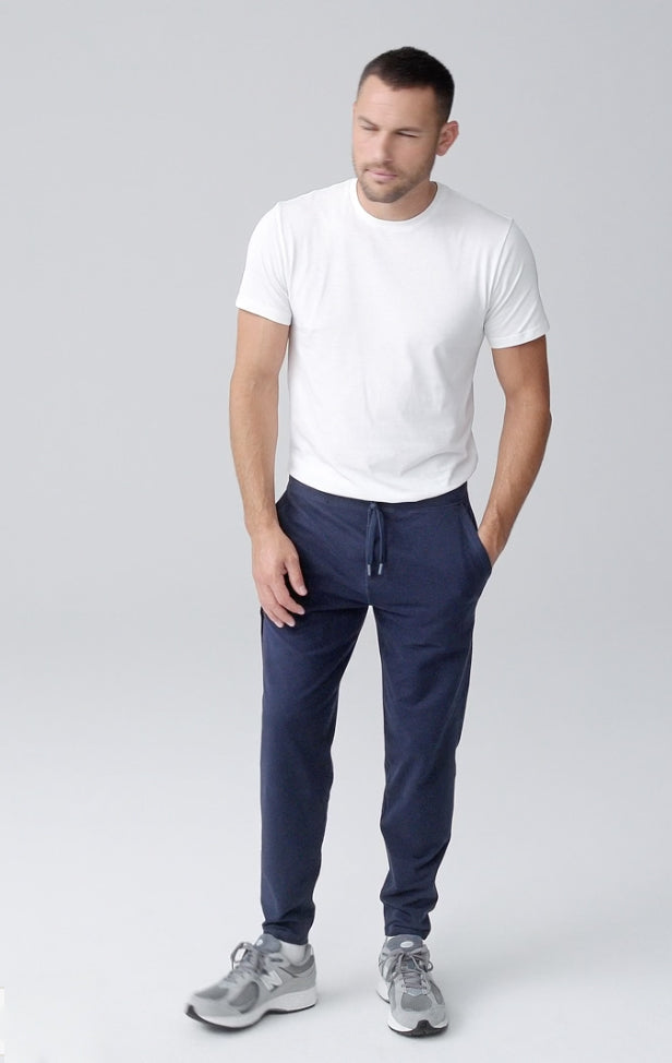 A man stands against a plain light gray background, wearing a white t-shirt, navy blue jogger pants, and gray sneakers. One hand is in his pocket and he is looking slightly downward.