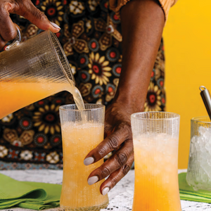 Person pouring orange juice into a glass with another glass of orange juice and ice cubes on a yellow background.