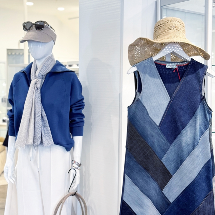 A store display shows a mannequin wearing a blue sweater, white skirt, beige cap, sunglasses, and scarf. Next to it hangs a sleeveless denim patchwork dress with a straw hat above it.