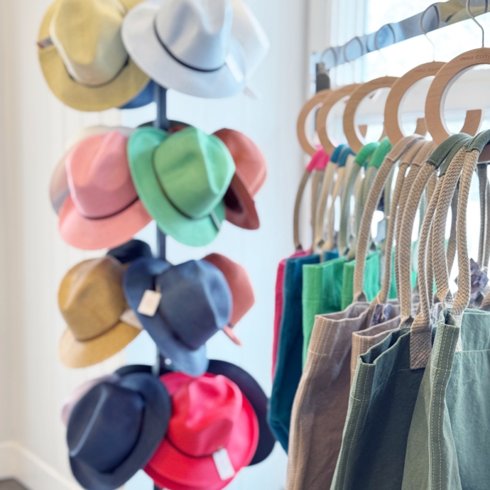 A display stand holds colorful hats, including pink, blue, green, tan, and red, next to a rack of reusable tote bags in various shades of green and blue, inside a well-lit shop.