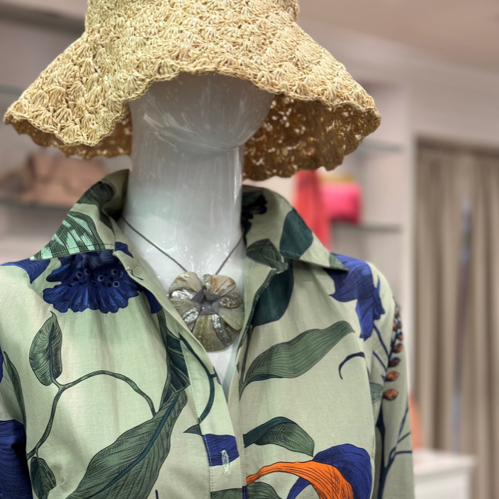 A mannequin wearing a straw hat, a floral-patterned button-up shirt with large green leaves, dark blue flowers, and orange accents, and a necklace with a large, flower-shaped pendant in a boutique setting.