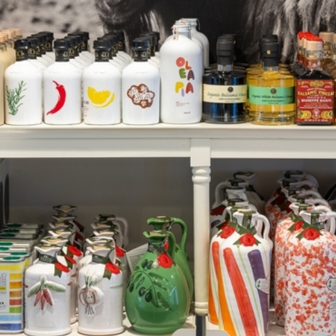 Assorted bottles of olive oil and vinegar with colorful, artistic labels and unique ceramic designs are neatly arranged on two white shelves in a shop.